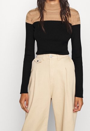 Frau trägt einen gerippten, langärmeligen Farbblock-Pullover in Beige und Schwarz, kombiniert mit hoch taillierter, weiter beiner Hose in Beige.