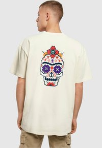 Kremfarget bomull T-skjorte med et levende pixel-kunst skull-design med blomsterdetaljer i rødt, lilla og blått på baksiden.