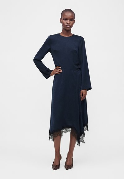 Femme portant une robe bleu marine à manches longues avec bordure en dentelle et ourlet asymétrique, debout avec une main sur la hanche et portant des talons foncés.