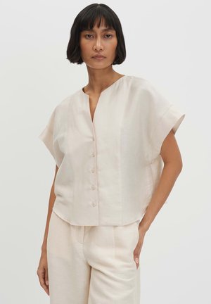 Femme aux cheveux courts noirs portant un haut crème ample à manches courtes boutonné et un pantalon large assorti, debout, une main dans la poche.