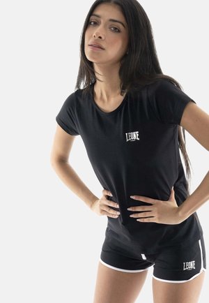 Femme aux longs cheveux foncés, debout les mains sur les hanches, portant un t-shirt noir Leone 1947 et un short assorti avec un liseré blanc.