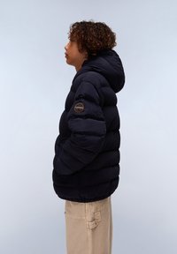 Černá péřová bunda s kapucí, se zaobleným logem na rukávu, prošívaným designem a elastickými manžetami. Nošena s béžovými kalhotami.