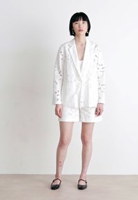 White textured blazer with lace cut-outs, paired with matching white shorts. Black flats complete the outfit, featuring a simple, elegant design.
