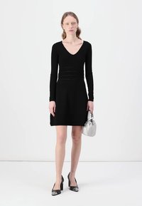 Black long-sleeve dress with a V-neck, textured knit design, fitted waist, and flared skirt. Paired with a silver handbag and black heels.