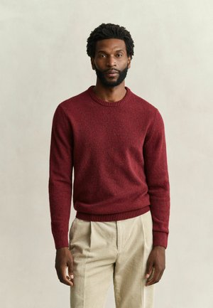 Homme aux cheveux courts et bouclés avec une barbe, portant un pull rouge en tricot et un pantalon beige en velours côtelé, debout devant un fond neutre.