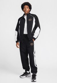 Chándal atlético negro con acentos blancos, que incluye una chaqueta con cremallera y pantalones de cintura elástica. El estilo de los zapatos es deportivo con una parte superior texturizada.