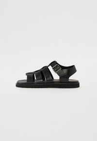 Black leather sandals with interwoven straps, a side buckle for adjustment, flat sole, and textured tread for grip.