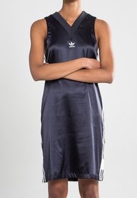 Osoba ubrana w bezrękawnikową, granatową sukienkę Adidas z białymi paskami po bokach, z założonymi rękami, na jednolitym tle.