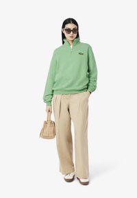 Sudadera verde con cremallera y logo bordado, pantalones anchos beige, y un pequeño bolso beige, complementados con zapatillas blancas y gafas de sol de gran tamaño.