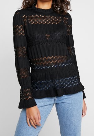 Black long-sleeve top with a ribbed texture, featuring wavy lace cutouts, a round neckline, and flared cuffs. Paired with light blue jeans.