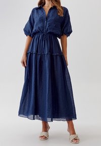 Mörkblå, lager-på-lager maxi klänning med korta, puffade ärmar, knappning fram, dragsko i midjan och rynkad textur, i kombination med flätade sandaler.