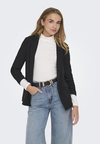 Črn blazer z natančnimi reverji, oblečen preko belega visokega ovratnika. Svetlo modri široki kavbojki in črn pas z zlatim sponko dopolnjujejo oblačilo.