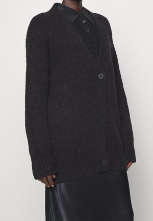Personne portant un cardigan en tricot texturé noir avec de gros boutons par-dessus une chemise noire à col et une jupe noire.