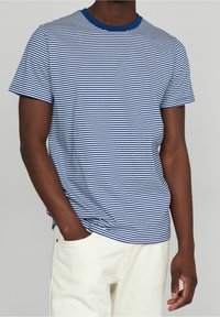 Gestreiftes T-Shirt mit blauen und weißen Streifen und kurzem Arm, mit einem blauen Rundhalsausschnitt, aus weichem Stoff, getragen mit cremefarbenen Hosen.