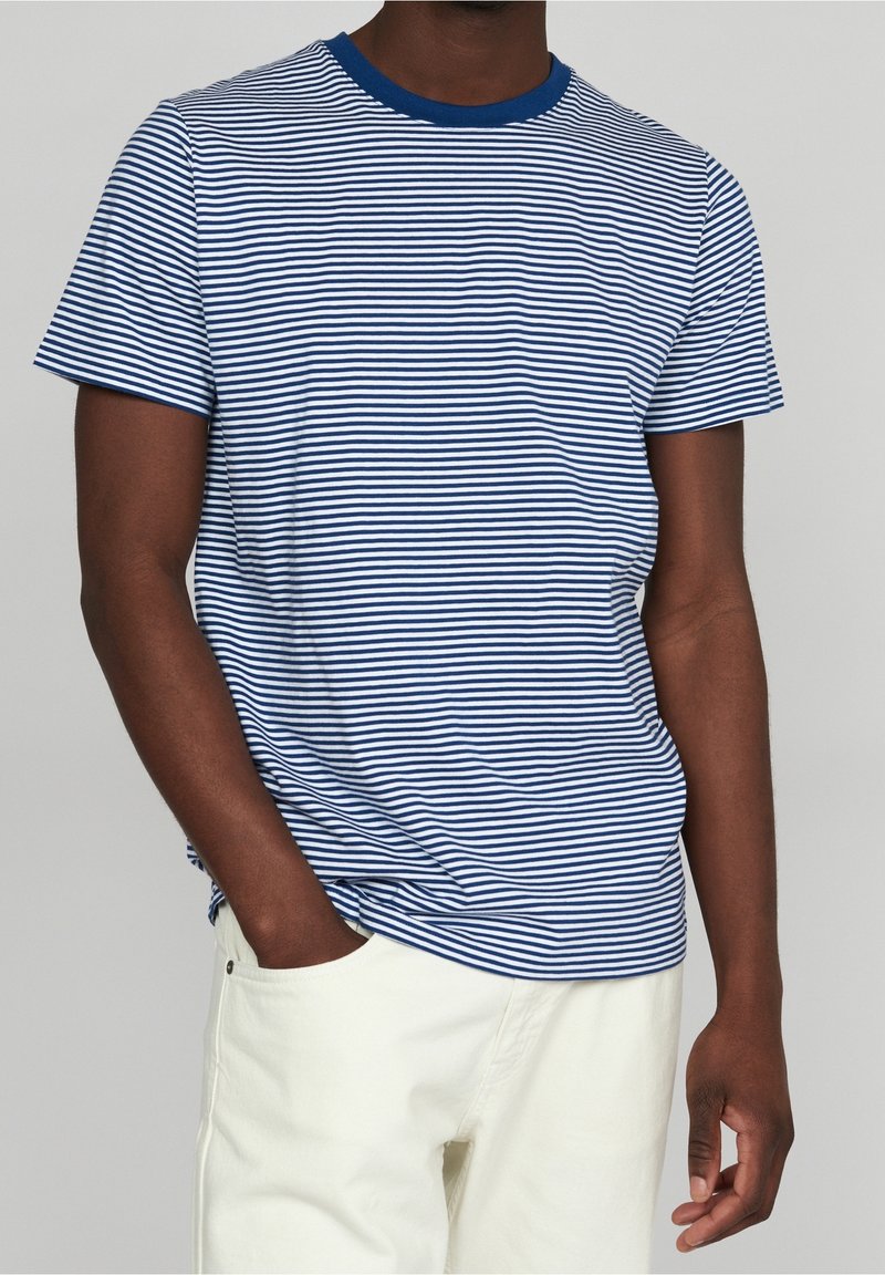 Gestreiftes T-Shirt mit blauen und weißen Streifen und kurzem Arm, mit einem blauen Rundhalsausschnitt, aus weichem Stoff, getragen mit cremefarbenen Hosen.