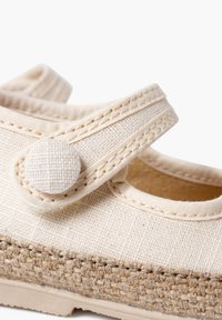 Light beige canvas shoe with a rounded toe, ankle strap, and textured jute sole. Features a fabric button for strap closure.