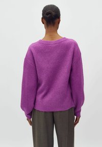 Frau mit dunkler Haut und hochgestecktem Haar trägt einen lila gerippten Pullover und graue Hosen, steht mit dem Rücken zum Betrachter vor einem einfachen weißen Hintergrund.