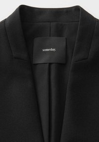 Schwarzer Blazer aus einem glatten Stoff, mit Reverskragen und einem Etikett, auf dem "irgendwann" steht. Das Innere ist schick und unauffällig.