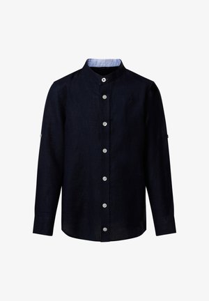 Garçon debout portant une chemise marine à boutons, un short cargo beige, des baskets blanches et un pull orange posé sur ses épaules.