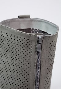 Bottine en daim gris avec motif perforé, laissant apparaître une doublure intérieure sombre. Dispose d'une fermeture éclair latérale et d'un bord supérieur arrondi. Texture lisse.