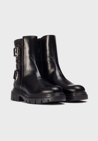 Botas de cuero negro hasta el tobillo con suelas de goma gruesas, cremalleras laterales, paneles traseros acolchados y dos hebillas plateadas en cada bota.