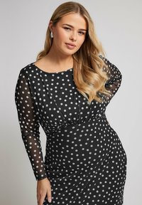 Musta pilkullinen mekko pitkien läpinäkyvien hihojen, rypytetyn yläosan ja istuvan siluetin kanssa, jossa on kontrastina valkoisia pisteitä ja pehmeä rakenne.