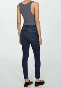 Jean skinny bleu foncé à taille haute, avec deux poches arrière et des détails de couture visibles, associé à un haut sans manches gris.