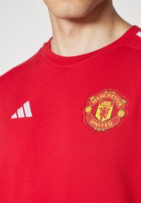 Primo piano di una persona che indossa una maglietta rossa del Manchester United con lo stemma del club e il logo bianco di Adidas sul petto.