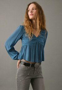 Blusa lunga teal a maniche lunghe con fronte arricciato e scollatura a V, abbinata a jeans grigi con cuciture decorative e una cintura nera con fibbia circolare.