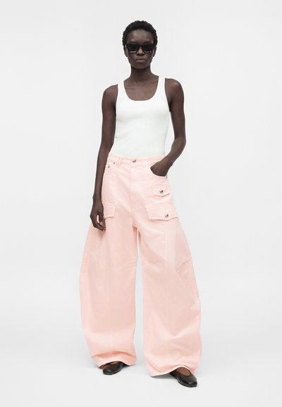 Ψηλή γυναίκα που φοράει λευκή αμάνικη μπλούζα, ανοιχτό ροζ φαρδιά παντελόνια cargo, μαύρα παπούτσια και μεγάλα μαύρα γυαλιά ηλίου, όρθια με το ένα χέρι στην τσέπη.