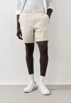 Mand iført beige elastiske shorts, hvid ribbet skjorte, hvide sneakers og hvide sokker, står op ad en almindelig væg.