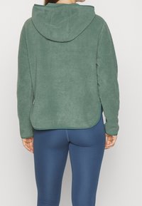 Grøn fleece-hoodie med en tekstureret overflade, med hætte, lange ærmer og en buet kant. Båret over blå leggings.
