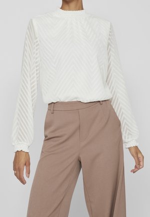 Vrouw die een witte textuur blouse met lange mouwen draagt, met chevronpatronen en een bruisbroek met hoge taille in bruin.