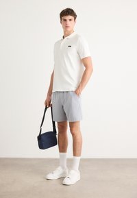 Hvit polo skjorte, lysegrå shorts, hvite sneakers og knelange sokker. Holder en liten marineblå crossover-veske. Enkel, uformell design.