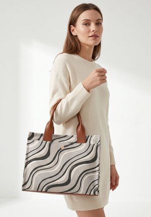 Donna con vestito maglione color crema che tiene una borsa tote beige e nera con motivo ondulato e manici in pelle marrone su sfondo bianco.