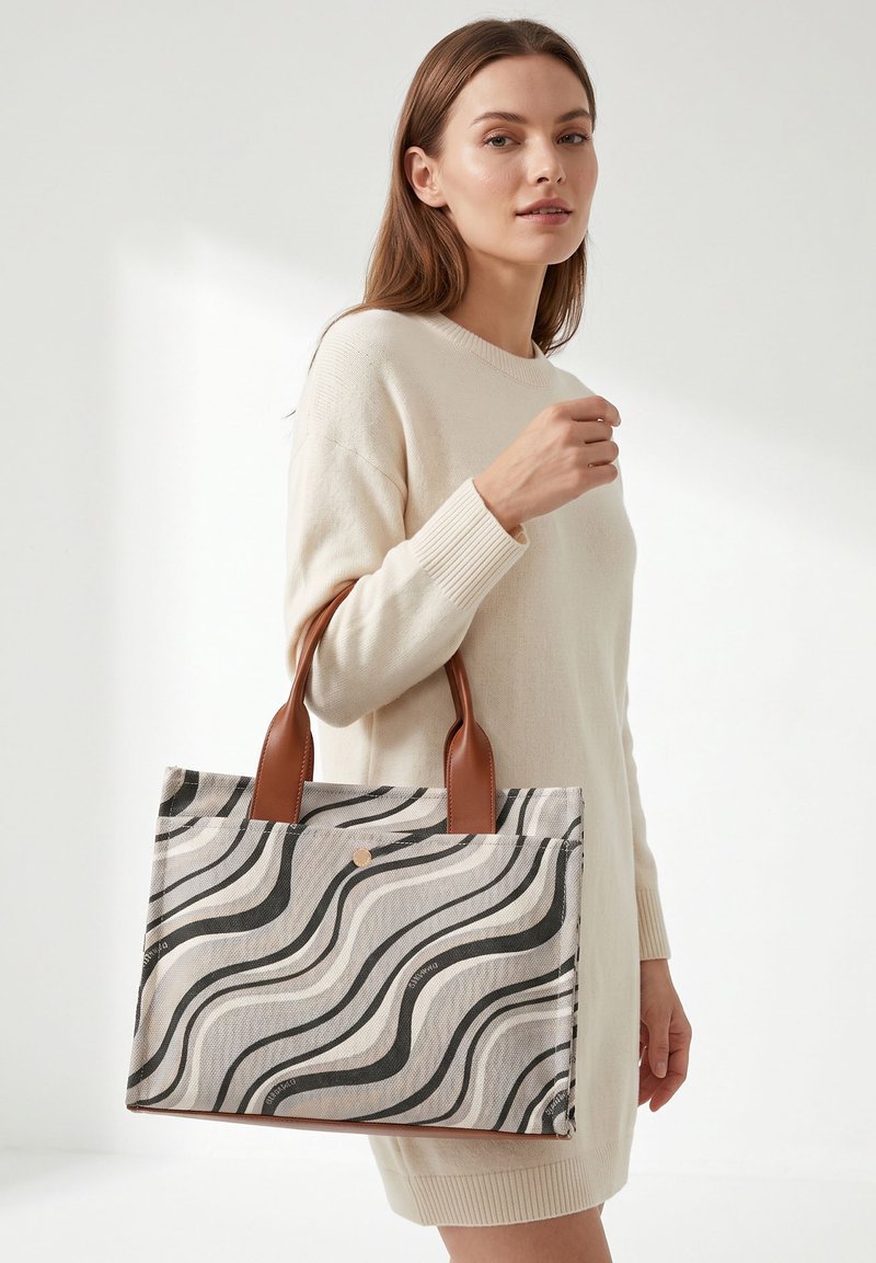Femme en robe-pull crème tenant un sac cabas beige et noir à motif ondulé avec des poignées en cuir marron sur fond blanc.