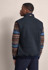 Dunkelblaues gepolstertes Westchen mit gemusterten Strickärmeln in Braun-, Blau- und Cremetönen. Hoher Kragen und gerippter Saum betonen die Struktur.