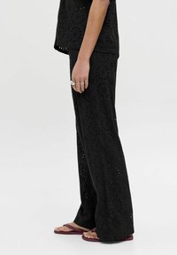 Pantalons en tissu noir avec un design évasé, présentant des motifs floraux découpés. Associés à des sandales plates simples.