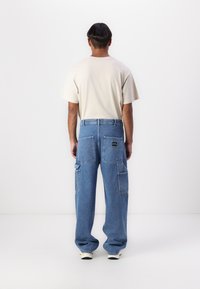 Light beige cotton t-shirt paired with loose blue denim cargo pants featuring multiple pockets and a logo patch on the back.