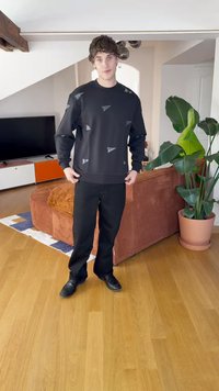 Felpa nera con motivi geometrici grigi, abbinata a pantaloni e scarpe neri. In piedi in una stanza ben illuminata con un divano e piante.