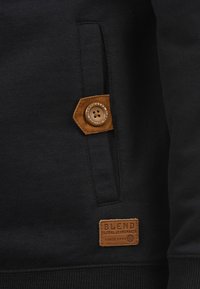 Schwarzer Sweatshirt mit strukturiertem Stoff, ausgestattet mit einem braunen Lederpatch und Knopfdetails. Beinhaltet eine seitliche Tasche für zusätzlichen Komfort.