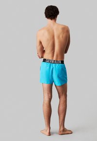 Costumi da bagno blu con un cinturino nero che presenta il marchio "CALVIN KLEIN". Materiale leggero. Progettati con una tasca sul retro.