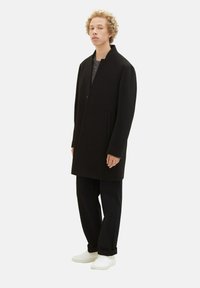 Schwarzer Mantel mit hohem Kragen, geradem Schnitt und zwei Seitentaschen. Über einem dunklen Oberteil getragen und kombiniert mit lockeren schwarzen Hosen und weißen Schuhen.