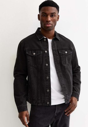 Man draagt een zwarte spijkerjas over een wit shirt, staande tegen een effen lichte achtergrond, handen ontspannen langs het lichaam.