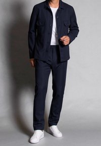 Navy fabric jacket with two front pockets, paired with matching trousers. White crew-neck T-shirt and white sneakers complete the outfit.