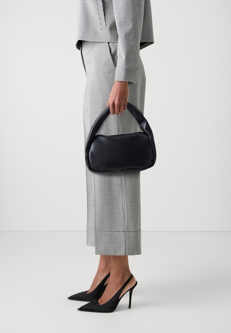 Femme en tenue à carreaux noir et blanc tenant un sac à main noir, portant des chaussures à talons hauts slingback noires, debout devant un fond uni.