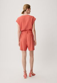 Koralle-texturiertes Blusenshirt mit kurzen Ärmeln, kombiniert mit korallenfarbener, hoch taillierter Shorts. Das Outfit hat einen Knoten an der Taille und Sandalen mit Blockabsatz.