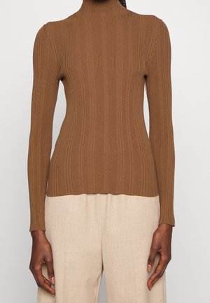 Personne portant un pull col roulé marron ajusté à côtes avec manches longues et pantalon ample beige clair sur un fond uni.