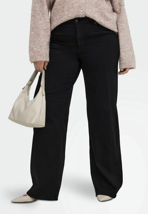 Personne portant un pantalon noir à jambes larges, un pull beige, des chaussures pointues beiges et tenant un petit sac à main beige dans la main gauche.