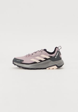 Zapatillas de trail con un upper ligero y transpirable de color púrpura claro, acentos en negro y una suela gris texturizada. Presentan tres rayas blancas y un tirador.